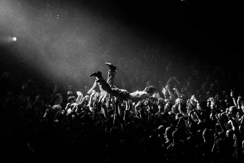 A person crowd surfs above a large audience at a concert, illuminated by a spotlight, while numerous hands reach up to support them. The image is in black and white.