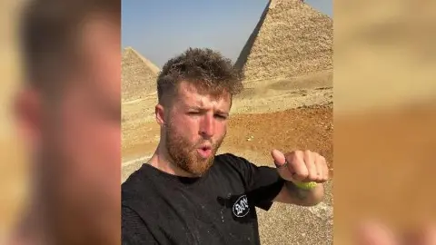 Tommy Greenhouse Tommy on a run in the desert with two pyramids in the background. He has short ginger hair and a beard and is doing a thumbs up sign at the camera.
