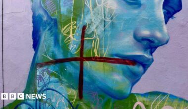 A dark red cross painted on top of a blue and green mural of a man looking down. It is painted on a purple wall, and there are several other blue and cream scribbles - which appear to be part of the mural - over his face.