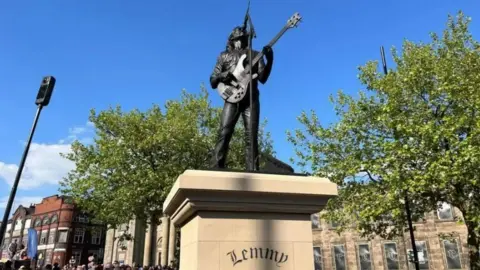 A bronze statue of Lemmy in his trademark pose, singing up into a mic while playing his bass guitar, which is pointing at one o'clock. There is a stone plinth below the statue with Lemmy engraved on it. There are trees and bright blue skies behind the statue.