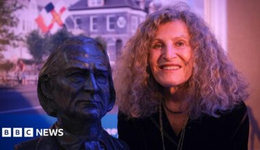 Nicole Farhi is posing next to the bust of Victor Hugo. She is smiling broadly and has shoulder-length curly hair. The bust shows Hugo looking serious. Behind them is a photo of the Victor Hugo centre which has a Guernseys flag flying in front of it.