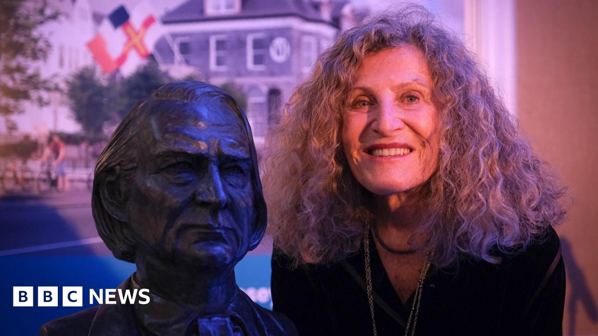 Nicole Farhi is posing next to the bust of Victor Hugo. She is smiling broadly and has shoulder-length curly hair. The bust shows Hugo looking serious. Behind them is a photo of the Victor Hugo centre which has a Guernseys flag flying in front of it.