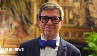 A close-up of Yann LeCun wearing thick rimmed black glasses, a navy suit with a dark blue bowtie standing in an opulent room at Buckingham Palace.