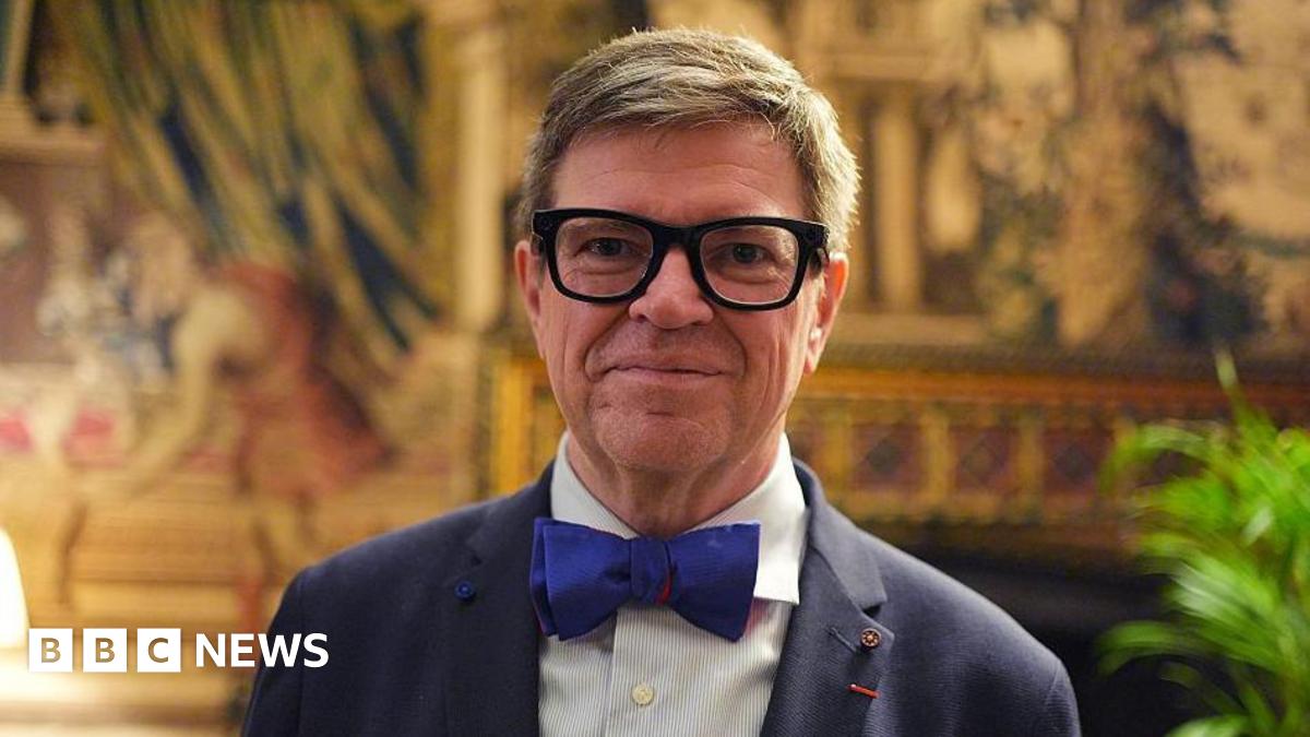 A close-up of Yann LeCun wearing thick rimmed black glasses, a navy suit with a dark blue bowtie standing in an opulent room at Buckingham Palace.
