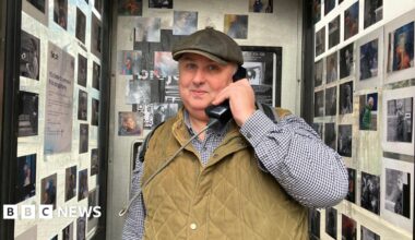 A man in a stone gilet, checked shirt and hat smiles as he holds a phone box phone to his ear. Behind him and to each sides are photos stuck onto the walls of the phone box.