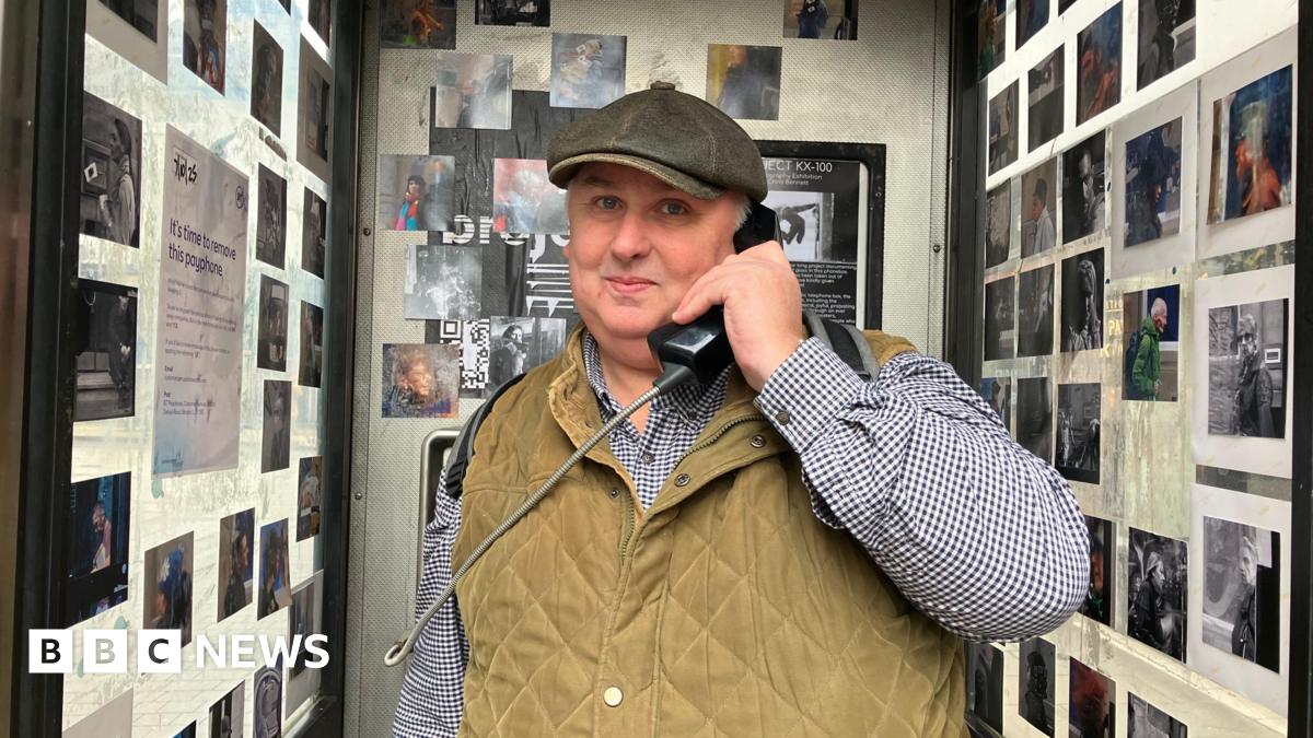A man in a stone gilet, checked shirt and hat smiles as he holds a phone box phone to his ear. Behind him and to each sides are photos stuck onto the walls of the phone box.