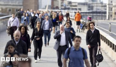 People in suits and casual outfits walk across a bridge on a sunny day.