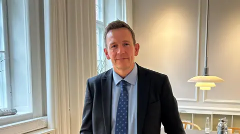 Rasmus Stoklund a middle aged man with short blonde hair wearing a suit and tie, next to a window in his office