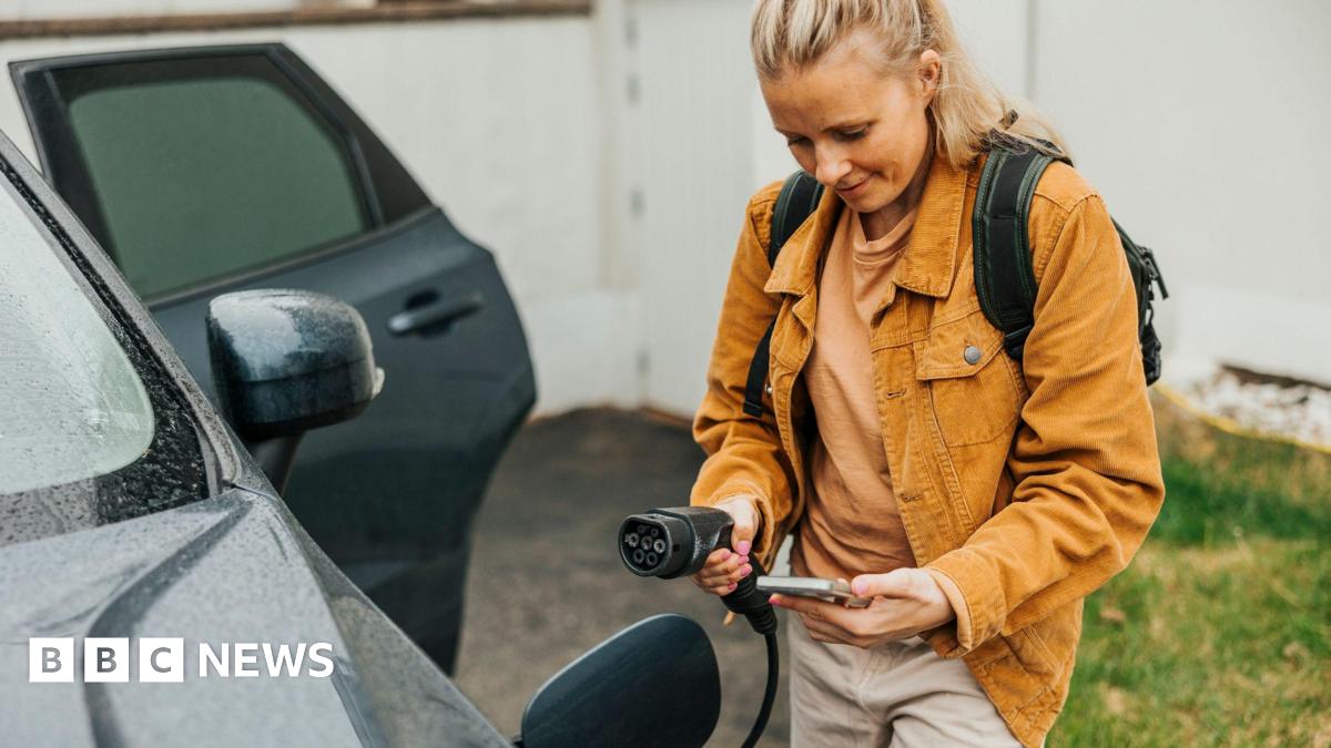 A young woman with her phone in her hand, about to plug an electrical charger into her car - she is wearing an orange jacket and has her blonde hair tied back into a pony tail, and is standing next to the car with the door opened