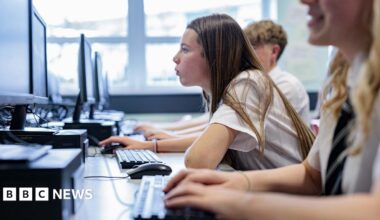 Profile of a teenage girl with long hair in school uniform in a classroom looking closely at a computer screen. Fellow students sit either side of her.