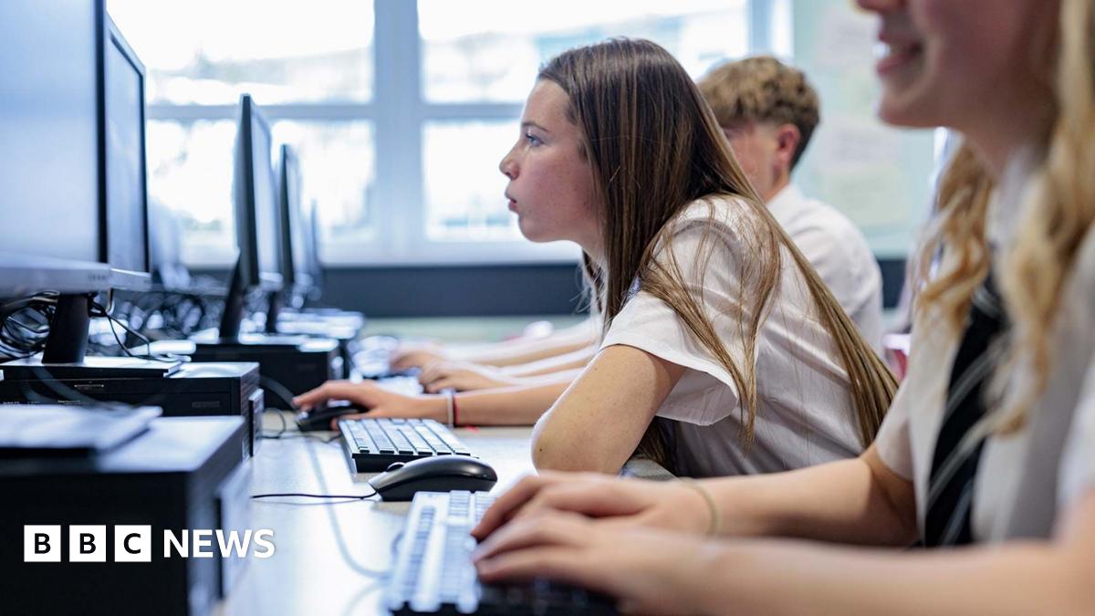 Profile of a teenage girl with long hair in school uniform in a classroom looking closely at a computer screen. Fellow students sit either side of her.