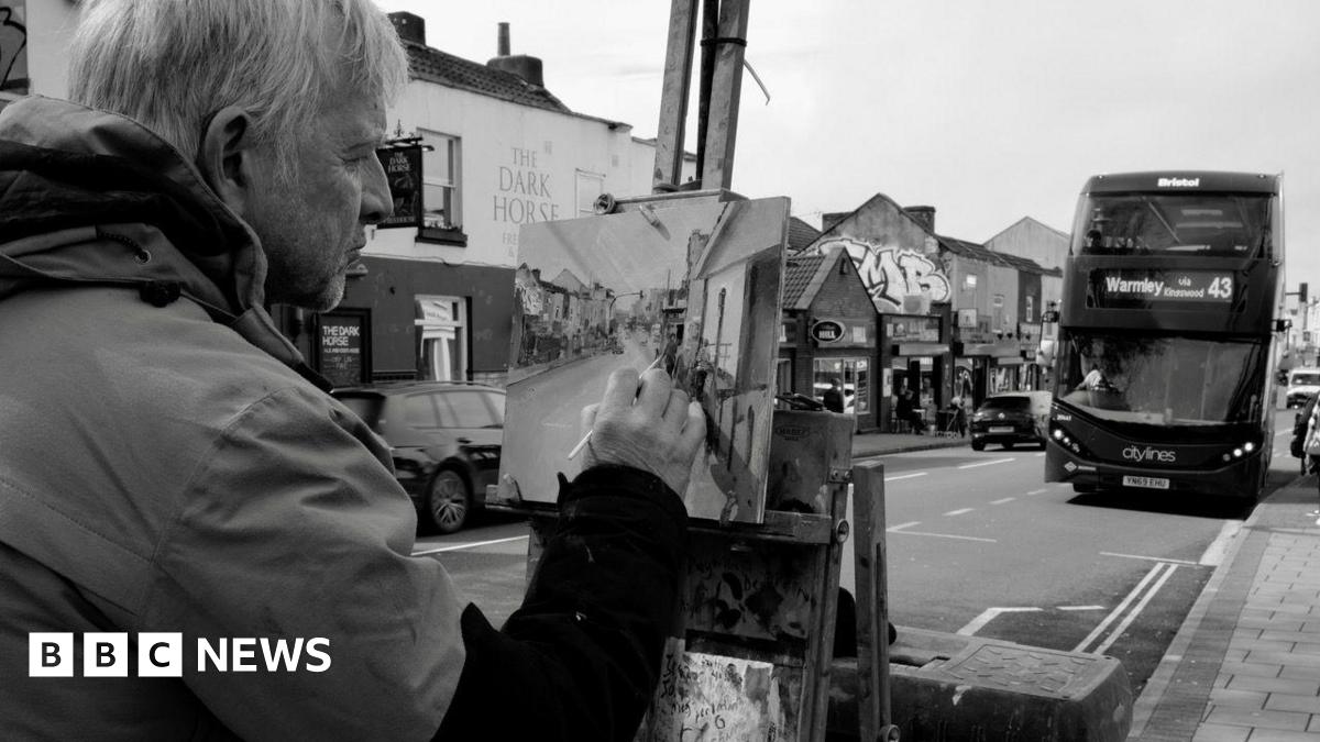 A black and white photo of artist Peter Brown painting a picture of a street. He is holding the paintbrush to the canvas and looking at his work. A bus heading for Warmley can be seen driving on the road ahead of him, and a pub and other businesses can be seen across the road. The photo is taken over Peter's shoulder, so only half his face can be seen.