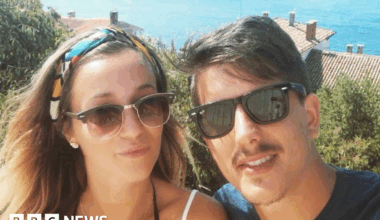 Young couple, one woman with medium brown hair and a young man, with short dark brown hair and a moustache both wearing sunglasses and looking at the camera taking a selfie. She has thin straps for a top and he is wearing a blue t-shirt - behind them are rooftops and the sea.