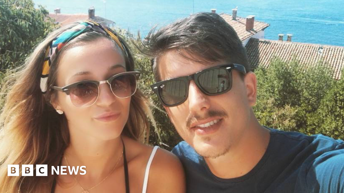 Young couple, one woman with medium brown hair and a young man, with short dark brown hair and a moustache both wearing sunglasses and looking at the camera taking a selfie. She has thin straps for a top and he is wearing a blue t-shirt - behind them are rooftops and the sea.