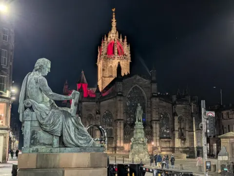 Alex Orr A grand Gothic-style cathedral dominates the scene, its ornate spire dramatically lit in red against the night sky. In the foreground, a bronze statue sits on a stone pedestal, overlooking the bustling street below. The area is illuminated by bright city lights, with people walking near the historic building.