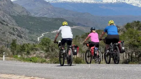 Craig Swan Craig, Sophie and Rona are on their bikes cycling a long a road which zig-zags up a mountainside in the distance. A mountain range rises in the distance.