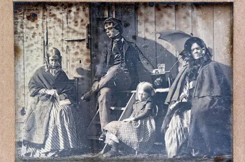 Private collection An early black and white image of four figures. Two women and a child are pictured in Victorian clothes, with large skirts, shawls and one is wearing a bonnet. A child wears a similar dress. A gentleman can be seen in a suit.