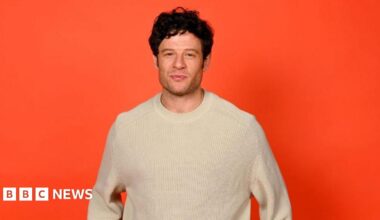 The actor James Norton wearing a white woollen jumper and standing in front of an orange back drop.
