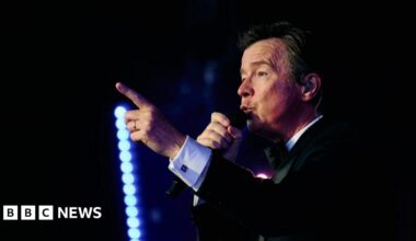 A man with dark hair and a dark suit in the dark, holding a microphone