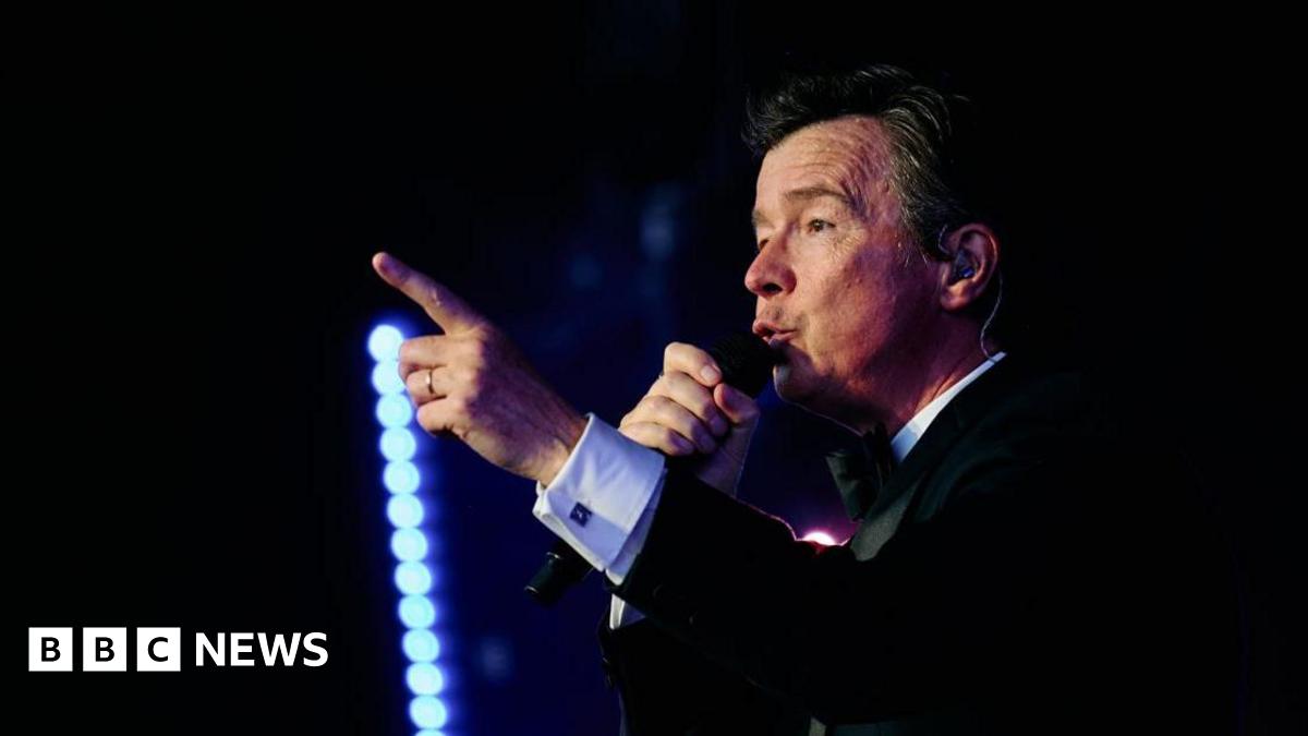 A man with dark hair and a dark suit in the dark, holding a microphone