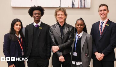 Sir Mick Jagger wearing a dark shirt and leather jacket. Four older children wearing school uniform and lanyards are stood either side of him. All are smiling at the camera.