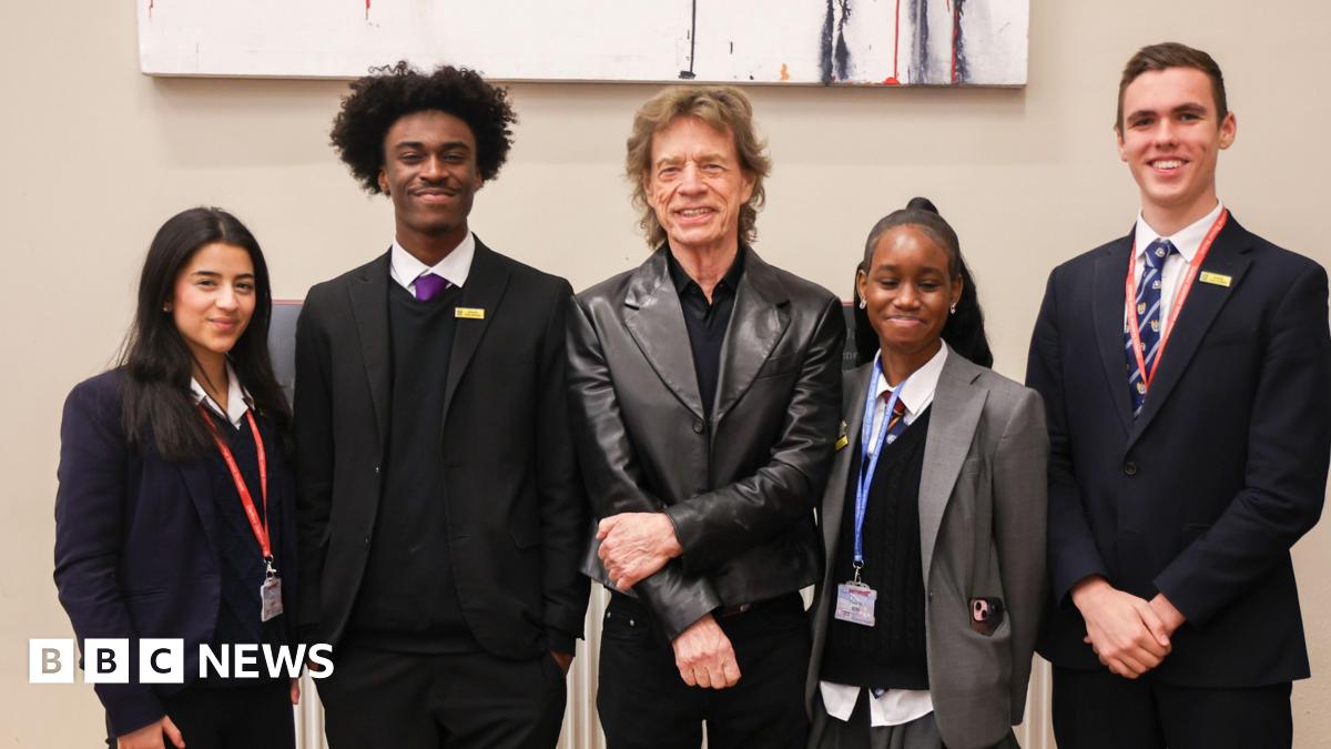 Sir Mick Jagger wearing a dark shirt and leather jacket. Four older children wearing school uniform and lanyards are stood either side of him. All are smiling at the camera.