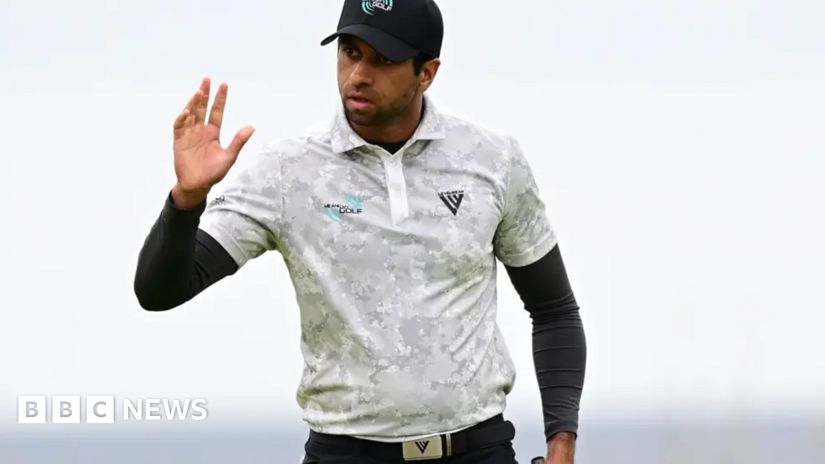 Rai is wearing golf gear and raising his right hand as a wave possibly to spectators. He has a white top on over a long sleeve black top and a white cap.