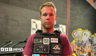 A man wearing a burgundy t-shirt and a black vest is pictured stood in front of a black painted wall.