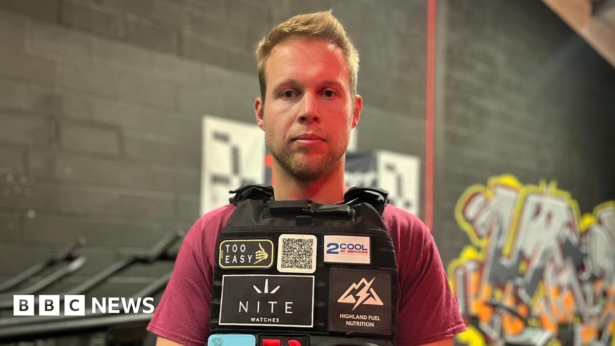 A man wearing a burgundy t-shirt and a black vest is pictured stood in front of a black painted wall.