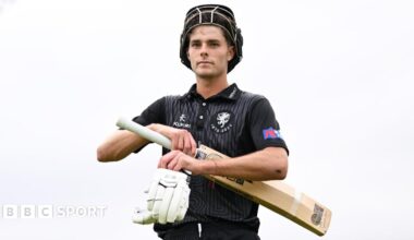 Archie Vaughan walking off the pitch with his helmet pulled up on his forehead and a his gloves and bat in his hands