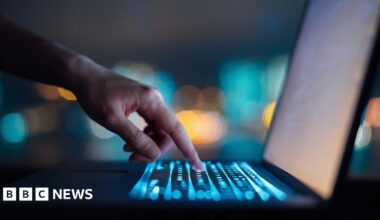 A man sits in front of a computer in the dark, with his silhouette illuminated by the light of the screen.