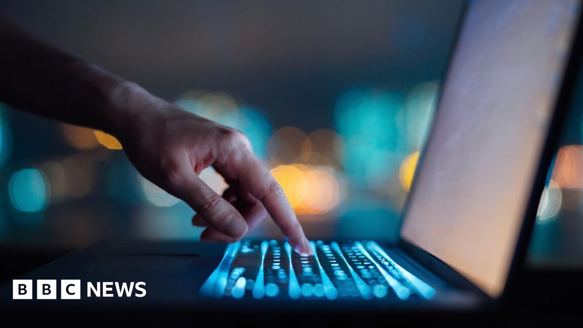 A man sits in front of a computer in the dark, with his silhouette illuminated by the light of the screen.