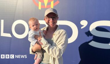 A woman wearing a cap and glasses stands in front of a sign at Lord's Cricket Ground in London, holding her infant son