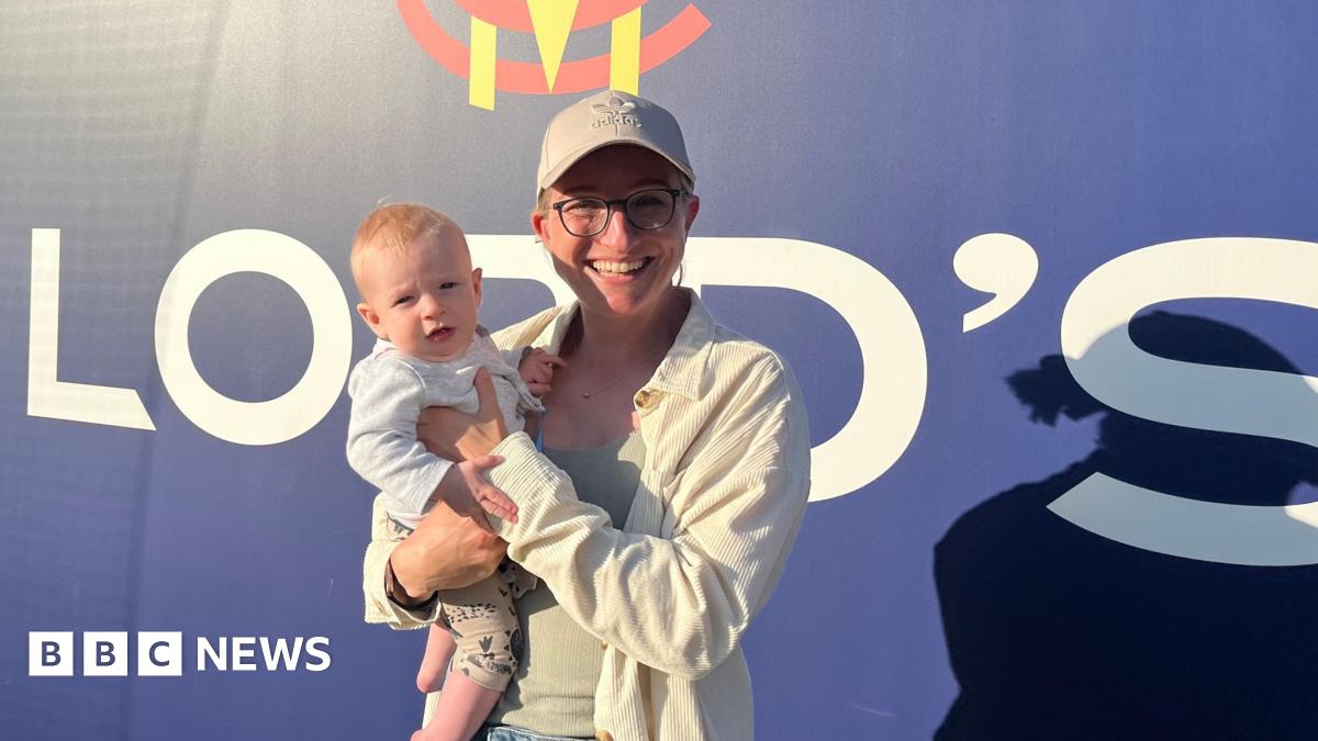 A woman wearing a cap and glasses stands in front of a sign at Lord's Cricket Ground in London, holding her infant son
