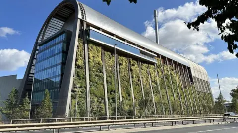 Leeds Civic Trust A large, modern building with a curved metallic roof. The front facade is covered with a living wall of plants. In the foreground there is a road with a metal barrier running through the centre of it.