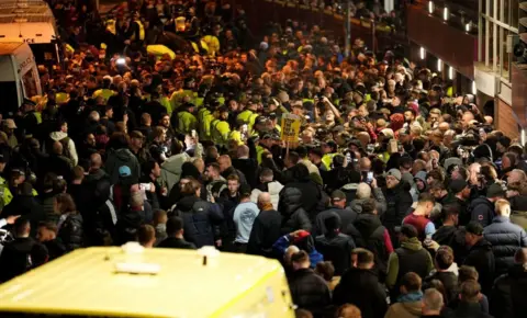 PA Media A large crowd gathers outside Villa Park. A police van is in the foreground, and police officers can be seen in the crowd, along with a number of banners.