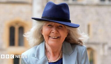 Jilly Cooper smiles at the camera. She has shoulder length grey hair and is wearing a light blue jacket and a dark blue top underneath, and a pearl necklace. She is also wearing a dark blue trilby-style hat