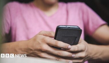 An unidentifiable person wearing a pink t-shirt holds and types on a smartphone in their hands.