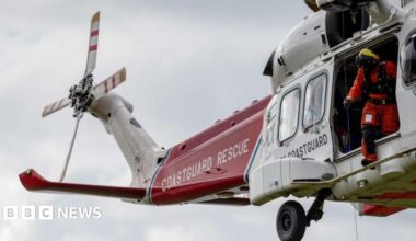 HM Coastguard helicopter crew conduct a search and rescue training exercise over the cliff tops in England. It is a red and white helicopter with signage reading 'COASTGUARD RESCUE' on it.