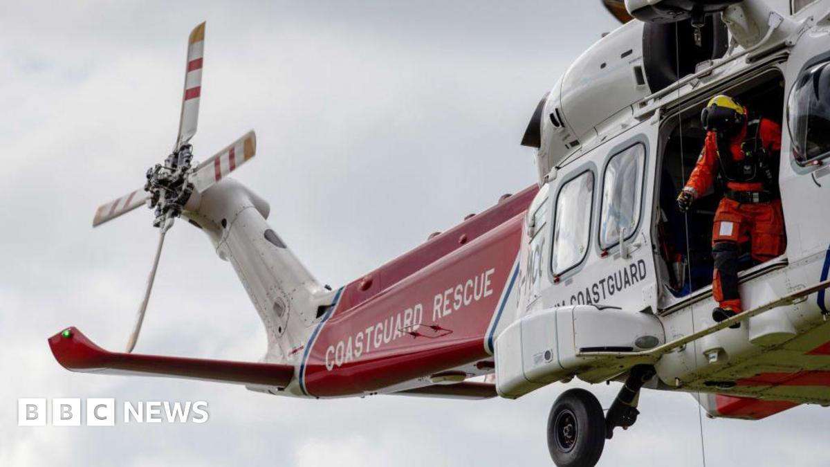 HM Coastguard helicopter crew conduct a search and rescue training exercise over the cliff tops in England. It is a red and white helicopter with signage reading 'COASTGUARD RESCUE' on it.