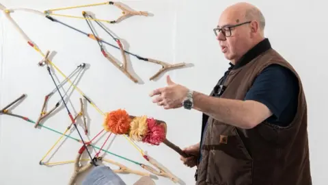 London Museum A man with black glasses and wearing a brown gilet holds a handmaid craft. In the background catapults are presented on the wall in odd directions. He holds a hand out and appears to be explaining something. 