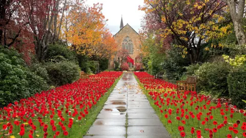 Norma Garthwaite A stone pathway leads to a historic church, flanked by vibrant autumn trees in shades of orange, yellow, and red. Both sides of the path are lined with hundreds of bright red poppies planted in green grass, creating a striking memorial display.
