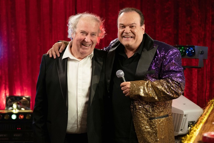 Shaun Williamson and Paul Bradley in EastEnders. They are both wearing suit jackets, with Williamson's being sparkly gold and purple. He is holding a microphone.