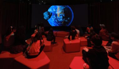 An Earth is projected on a dark screen as children sit on blocks in a dimly red-lit theater