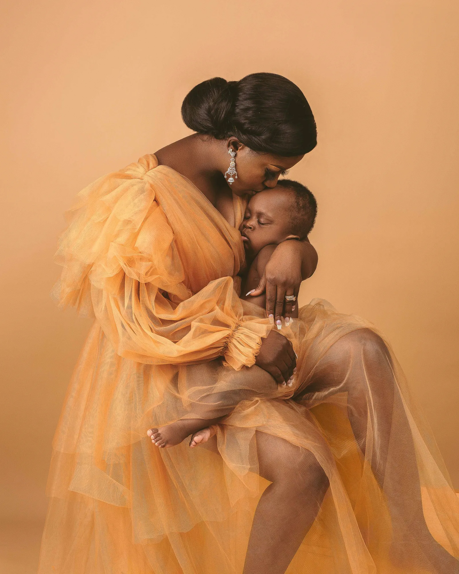 A mother in a flowing orange dress tenderly holds her baby, set against a warm, soft backdrop, exuding love and warmth
