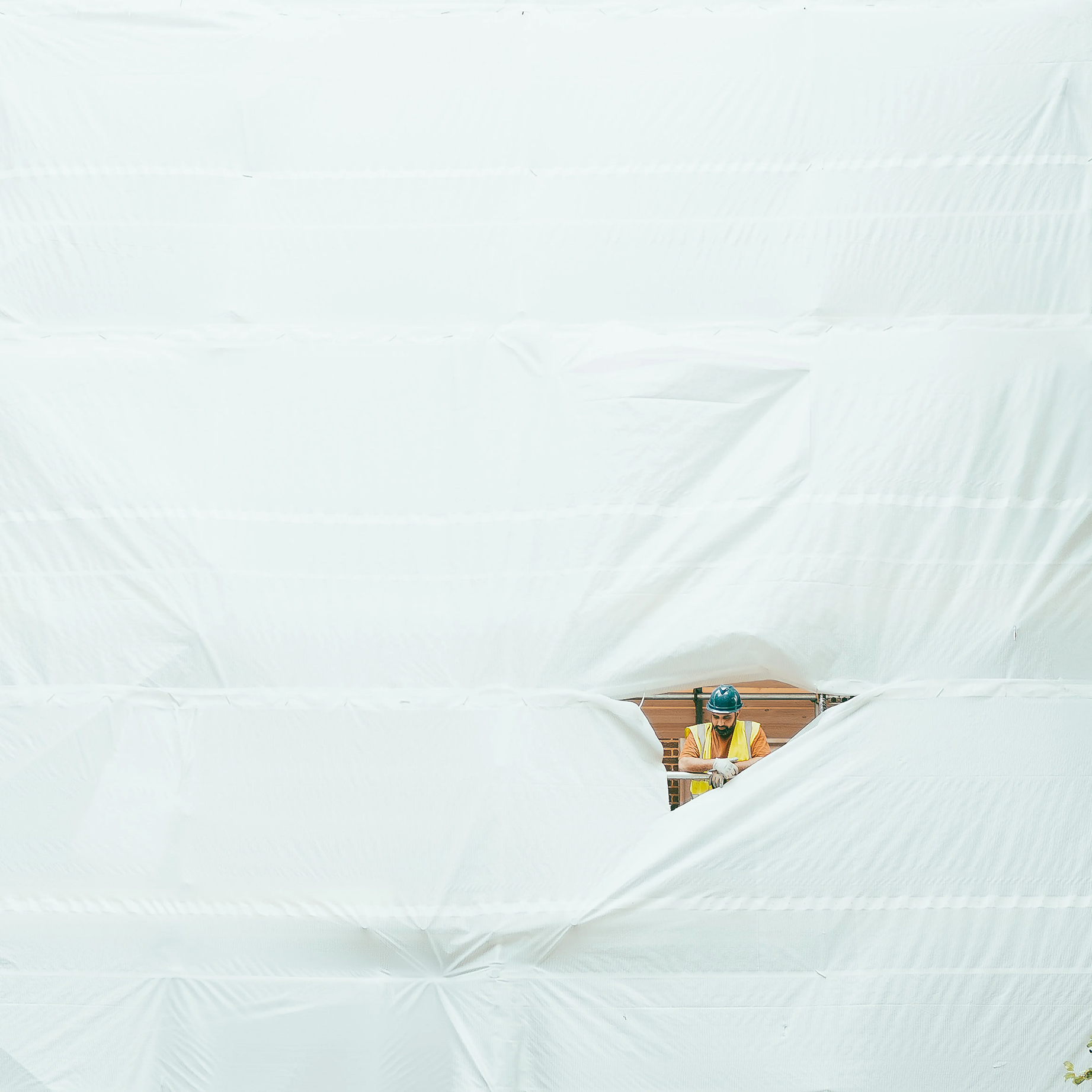 A construction worker in safety gear observes from behind a large white tarp, with a view framed by a tear in the fabric