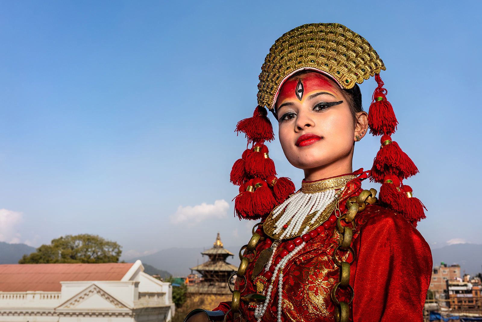 A figure in traditional attire with intricate red and gold garments, adorned with colorful tassels, against a clear blue sky