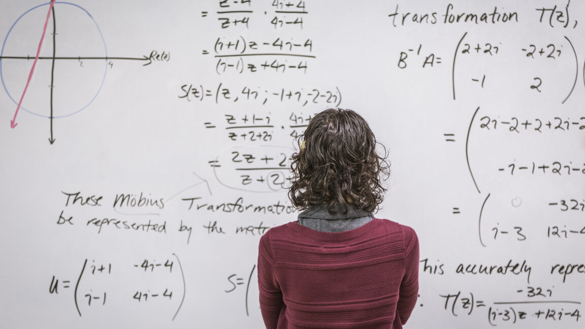 A woman standing in front of a white board that is packed with equations.