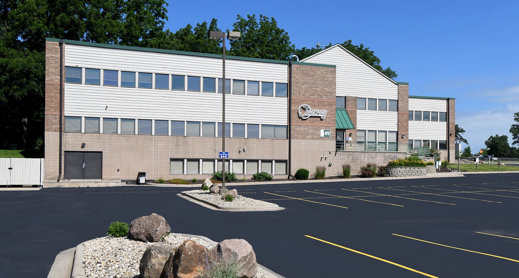 A two-story commercial building with a mostly empty parking lot, landscaping with large rocks, and a clear blue sky.
