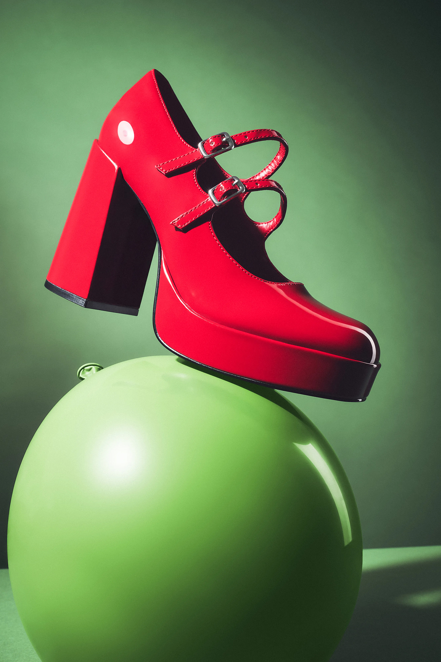 A vibrant red high-heeled shoe with two buckles perched atop a glossy green balloon against a soft green background